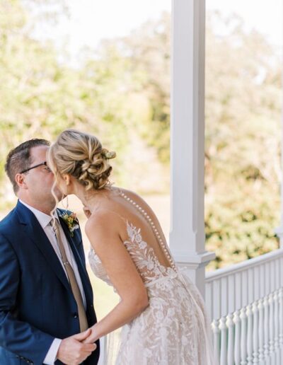 A couple kissing on a porch, with the woman wearing a lace wedding dress and the man wearing a blue suit. Trees and grass are visible in the background.