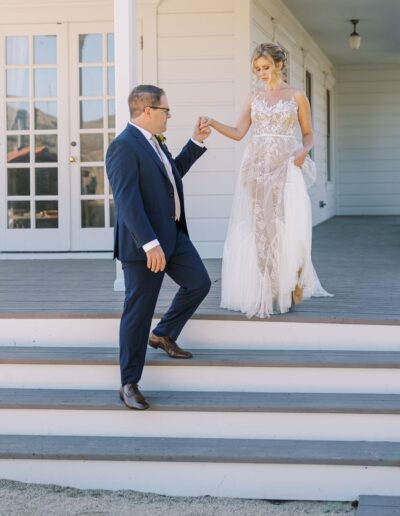 A man in a suit helps a woman in a lace wedding dress down a set of steps outside a white building.