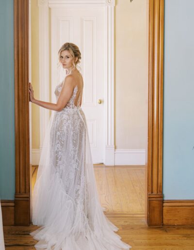 A woman in a white lace wedding dress stands in a doorway, looking back over her shoulder. She is in a room with wooden floors and a white door.