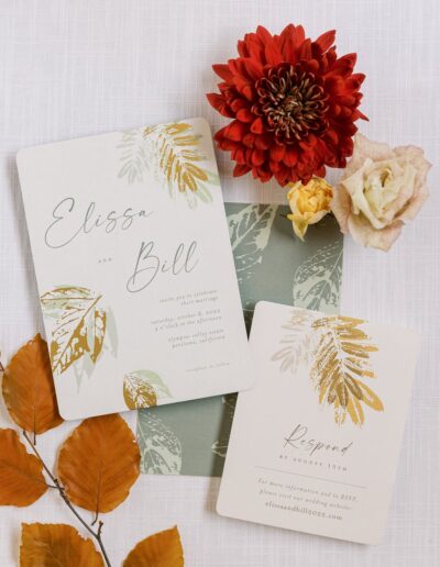 Wedding invitation suite with autumn leaf designs, featuring a large red dahlia, pale rose, and orange leaves. Text reads "Elissa and Bill" with RSVP details.