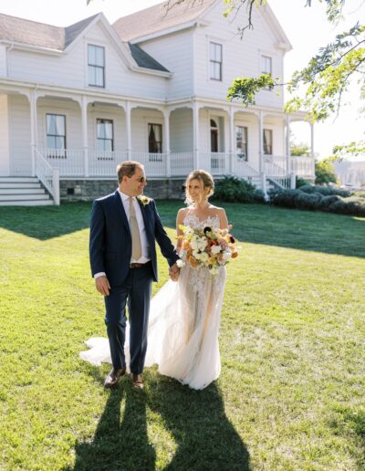 A couple walks hand in hand on a grassy lawn in front of a white house. The woman holds a bouquet of flowers.