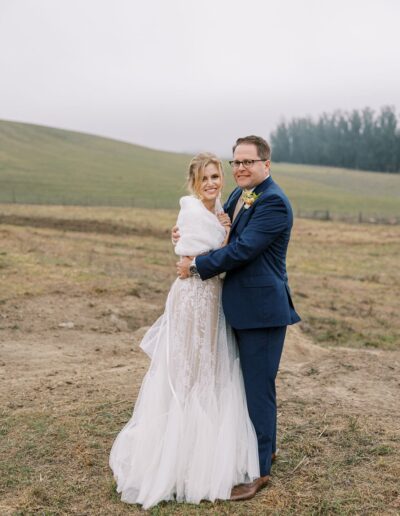 A bride and groom stand in an outdoor field. She wears a white gown and shawl; he wears a blue suit. They smile while embracing, with a grassy hill and overcast sky in the background.