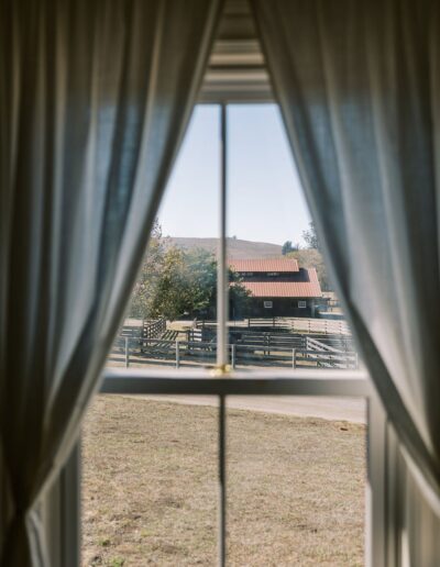 View of a rustic house with a red roof and wooden fence, seen through a window with sheer white curtains.