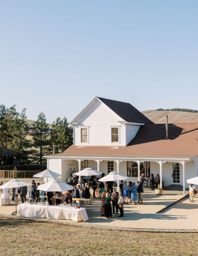 Outdoor gathering in front of a white house with attendees standing under umbrellas. Tables are set up with tablecloths and drinks. Trees and hills are in the background.