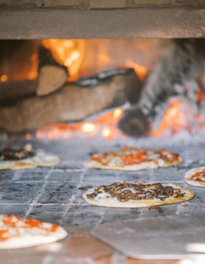 Pizzas baking in a wood-fired oven with visible flames and stacked logs in the background.