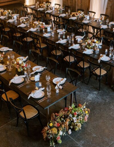 A long wooden table set with white plates, cutlery, and glassware. Chairs surround the table, and floral arrangements are placed at intervals, including one large display on the floor.