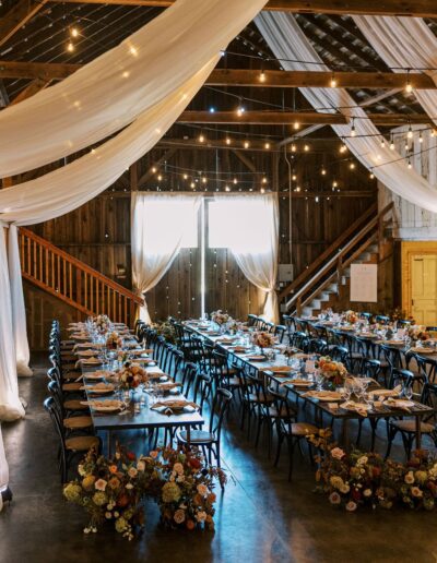 A rustic event hall with wooden beams and draped fabric, featuring long banquet tables set with tableware and floral centerpieces, illuminated by string lights.