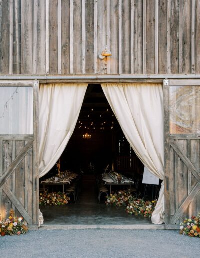 Entrance to a rustic barn with open wooden doors and white curtains, revealing a table set for an event. String lights and small floral arrangements line the entrance.