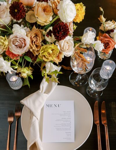 Elegant table setting with a menu card on a plate, surrounded by a floral centerpiece, wine glasses, and utensils. Roses and candles enhance the decor on a dark wooden table.