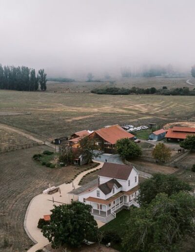 Aerial view of a farm with scattered buildings, fields, and trees under a cloudy sky.