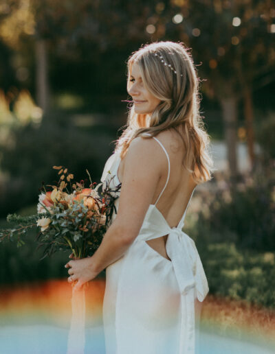 Woman in a white backless dress holds a bouquet of flowers outdoors, with sunlight creating a rainbow effect.