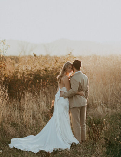 A bride and groom embrace in a field at sunset, with the bride wearing a flowing white dress and the groom in a beige suit.