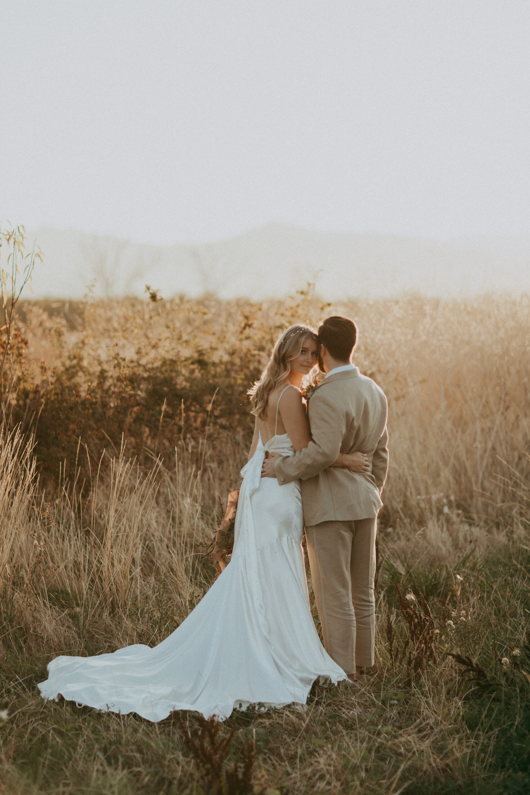 A bride and groom embrace in a field at sunset, with the bride wearing a flowing white dress and the groom in a beige suit.