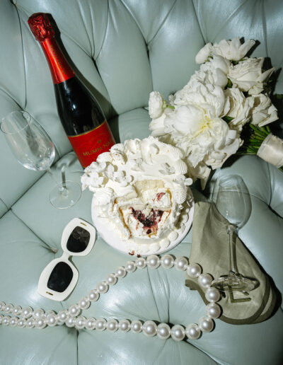 A cake slice, champagne bottle, two glasses, white flowers, sunglasses, and pearl necklace arranged on a light blue leather surface.