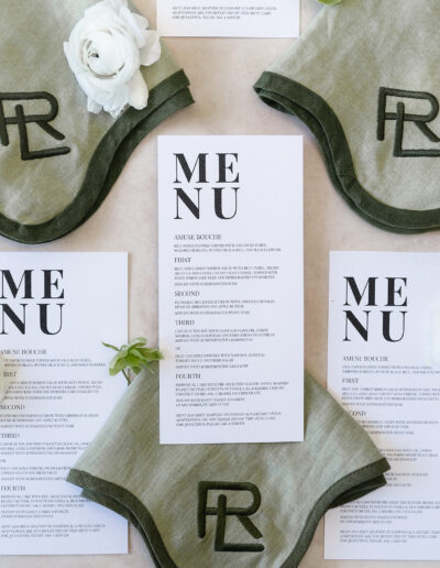 Restaurant menu placed on a table with green napkins embroidered with a logo and white flowers for decoration.