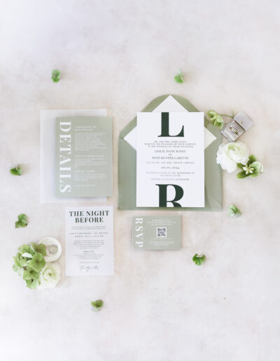 A flat lay of elegant wedding invitations and RSVPs in shades of green and white, surrounded by small flowers and a ring box on a light background.