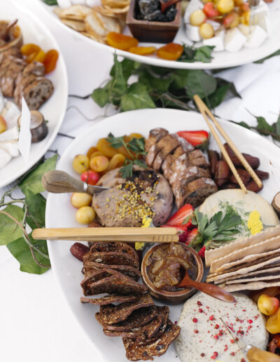 A charcuterie platter with various meats, cheeses, crackers, fruit, and dips, garnished with herbs and flowers on a white table.