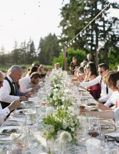 Outdoor dinner gathering with people seated at a long table decorated with white flowers, under string lights, and surrounded by trees.