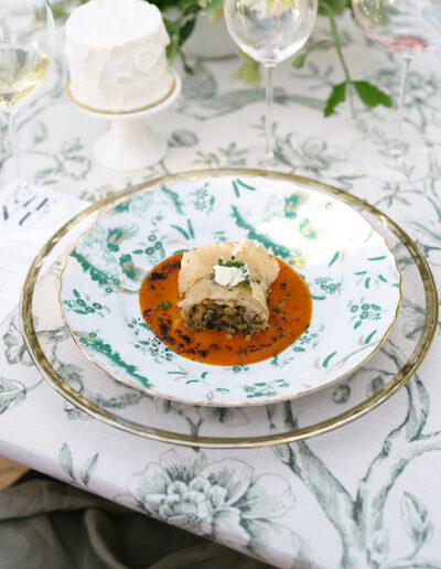 A gourmet dish of savory strudel on a patterned plate, garnished with a sauce and herbs, set on an elegant floral tablecloth with wine glasses and a small cake in the background.