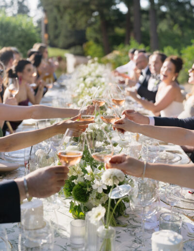 Guests clink glasses at a long, outdoor dining table adorned with flowers, celebrating in a lush garden setting.