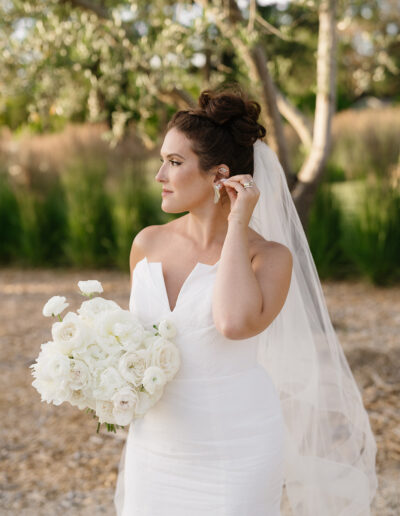 A bride in a white dress holds a bouquet of white flowers and adjusts her earring outdoors. Trees are visible in the background.