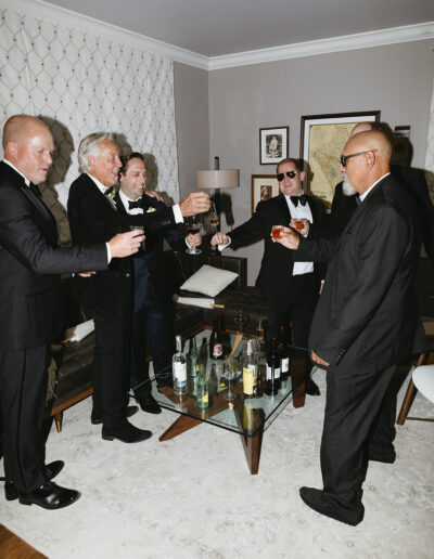 A group of men in suits stand in a living room holding drinks, toasting around a glass coffee table with bottles.