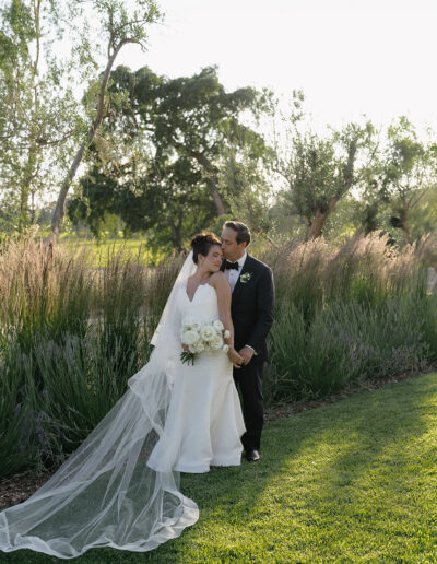 A bride and groom stand on a grassy lawn, with the groom kissing the bride's forehead. She holds a bouquet and wears a long veil. Trees and tall grasses are in the background.