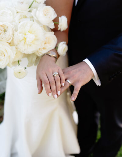 Bride and groom displaying wedding rings; bride holds a bouquet of white flowers.