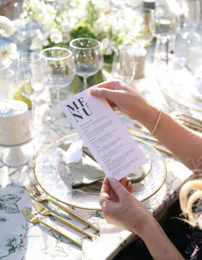 Person holds a white menu above a table set with elegant dinnerware and glassware, surrounded by floral decorations.