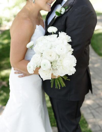 A bride in a white dress holds a bouquet of white flowers while standing next to a groom in a black tuxedo. They are outside on a paved path with grass in the background.