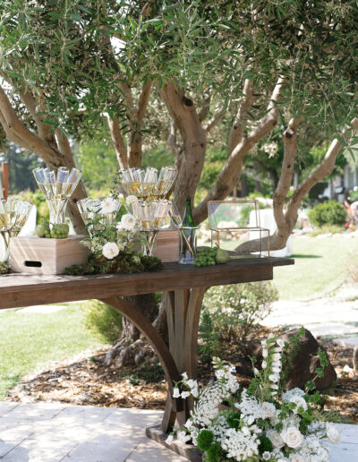 A rustic wooden table outdoors holds floral arrangements, champagne glasses, and decorative greenery, set against a backdrop of trees and grass.