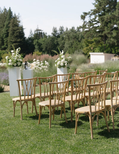 Wooden chairs arranged on grass facing floral arrangements, set outdoors with greenery and trees in the background, suggesting a wedding or event setup.