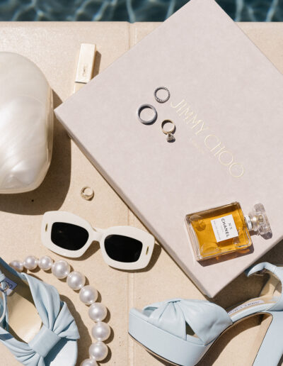 A pair of blue high heels, large pearl necklace, white sunglasses, Jimmy Choo box, Chanel perfume, rings, and a white clutch arranged neatly on a beige surface by a pool.