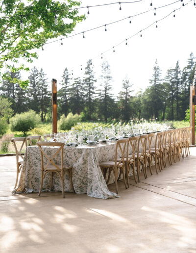 Outdoor dining setup with a long table, floral tablecloth, wooden chairs, and ambient string lights. Surrounded by trees and greenery.