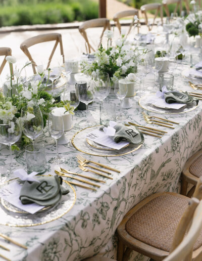 Elegant outdoor table setting with floral tablecloth, white flowers, and greenery centerpieces. Plates with green napkins and monograms; glassware and gold cutlery arranged neatly.