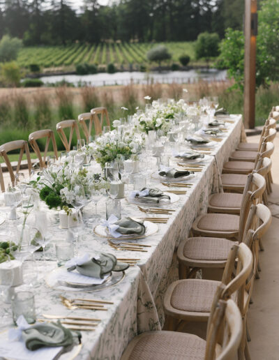 A long outdoor dining table is elegantly set with floral centerpieces, glassware, and neatly arranged tableware. Wooden chairs are lined up along both sides, with a view of a vineyard in the background.