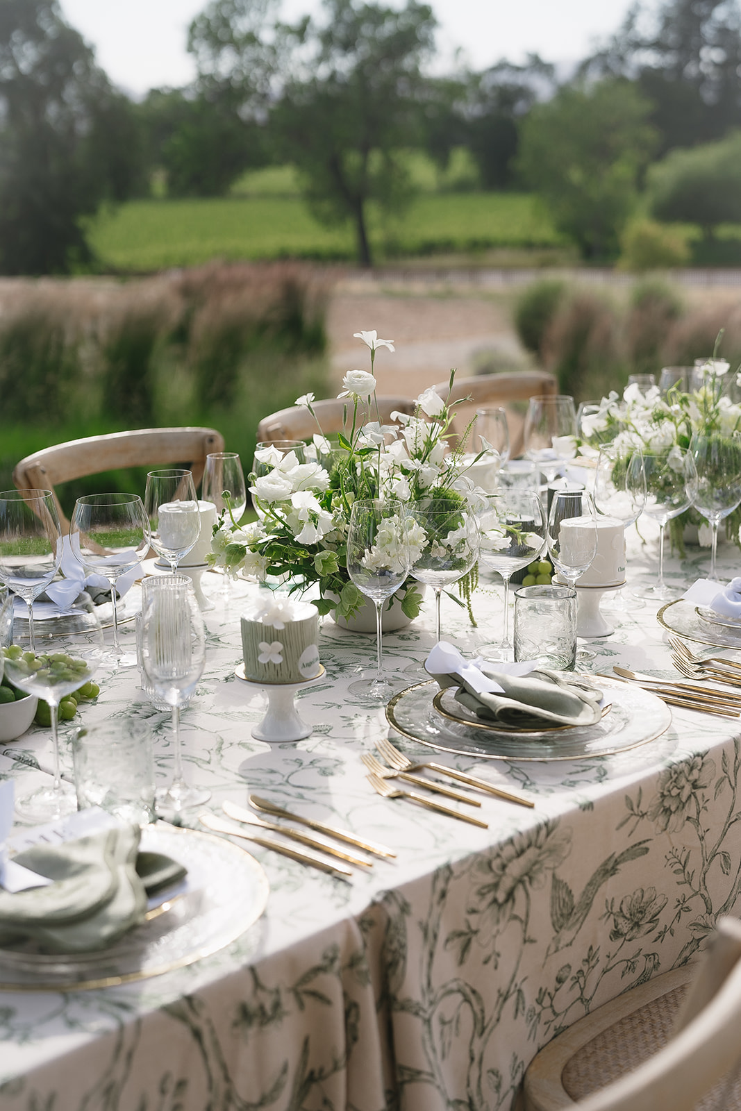 Elegantly set outdoor dining table with floral centerpieces, white tablecloth, glassware, and silverware, surrounded by greenery in the background.