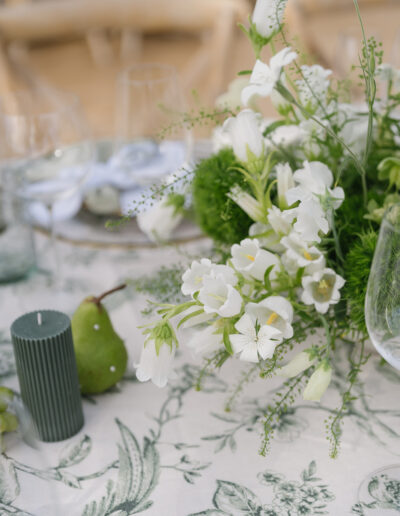 A floral table setting with white bell flowers, a green candle, and a pear on a patterned tablecloth.