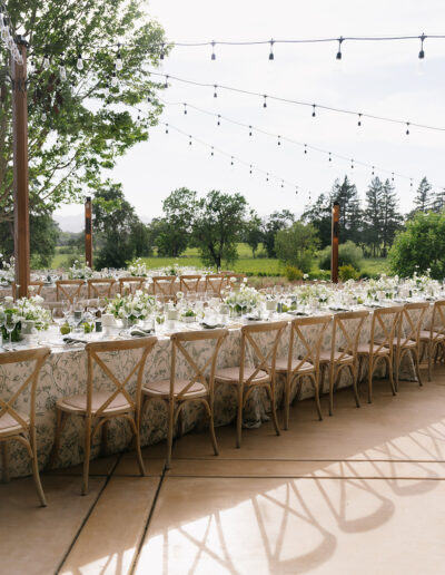 Outdoor dining setup with long tables covered in floral tablecloths, set with glassware and decorations. Wooden chairs surround the tables. String lights hang above.