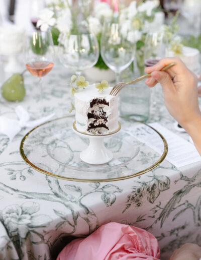 A person holding a fork near a partially eaten white cake on a plate. The table is covered with a floral-patterned cloth, surrounded by wine glasses and a floral arrangement.