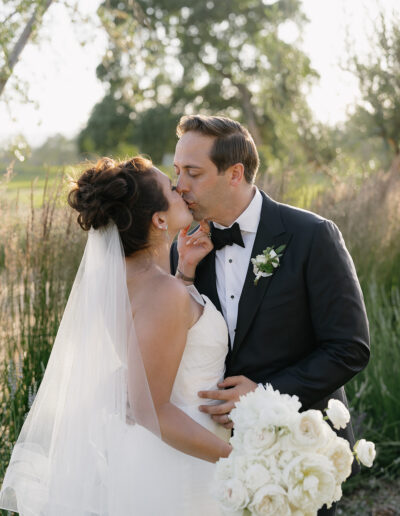 Bride and groom kissing in an outdoor setting, with the groom wearing a black suit and the bride in a white dress, holding a bouquet of white flowers.