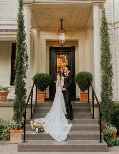 A couple in wedding attire stands on the steps of a large house with a lantern above, flanked by two tall trees. A bouquet rests on the steps.