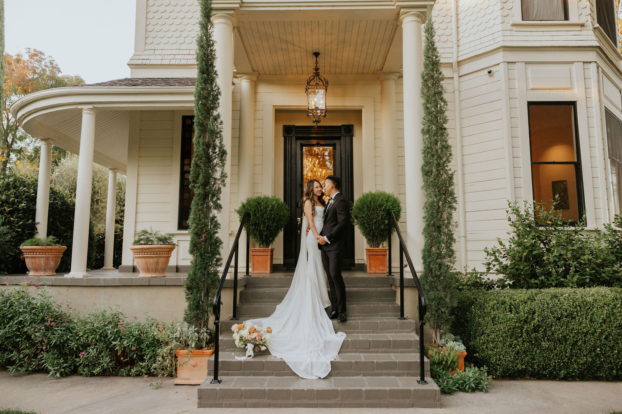 A couple in wedding attire stands on the steps of a large house with a lantern above, flanked by two tall trees. A bouquet rests on the steps.