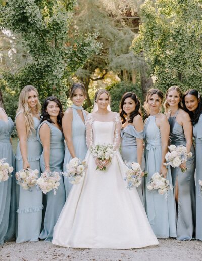 A bride stands with nine bridesmaids. The bride wears a white dress, and the bridesmaids wear matching light blue gowns. They are outdoors, surrounded by greenery.