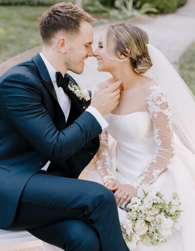 A bride and groom sit closely, smiling at each other. The bride wears a white dress with lace sleeves and a veil, holding a bouquet, while the groom is in a dark suit with a bow tie.