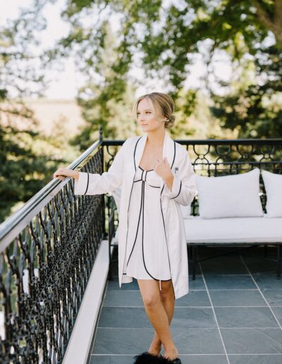 A woman in a white robe and black slippers stands on a balcony. She is looking to the side with trees in the background.