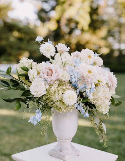 A white vase filled with a bouquet of white and pale pink roses, dahlias, and hydrangeas, accented with light blue flowers, set against a blurred green outdoor background.