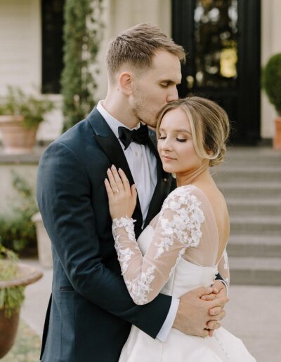 A groom in a black suit kisses the forehead of a bride in a white lace dress as they embrace outdoors on a sunny day.