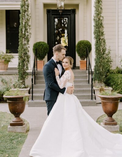 A couple in formal attire embrace in front of a white building with manicured shrubs and steps. The woman wears a white gown, and the man is in a dark suit.