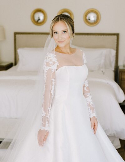 A bride in a white gown with lace sleeves stands in a bedroom, in front of a bed, smiling at the camera. Three circular mirrors are on the wall behind her.